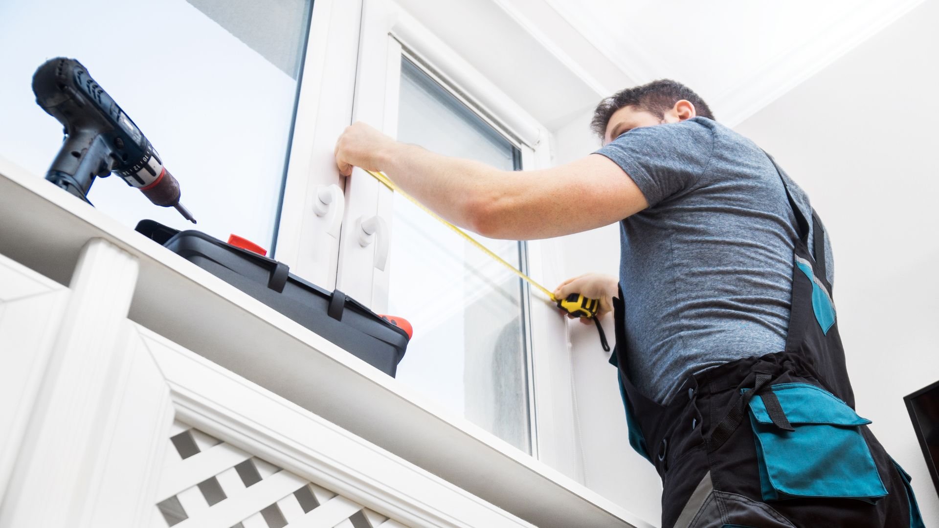Worker measuring window with tape measure and drill nearby